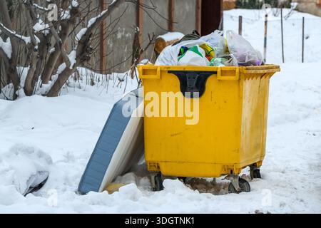 Overflowing yellow garbage container with trash bags in snowy winter yard, selective focus Stock Photo
