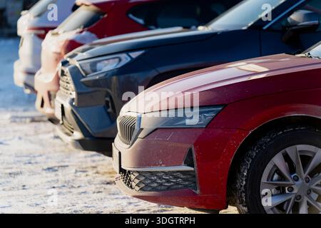 Parked cars aligned on frozen ground in winter, close view of red sedan and dark vehicles showing front bumpers wheels and headlights outdoors Stock Photo