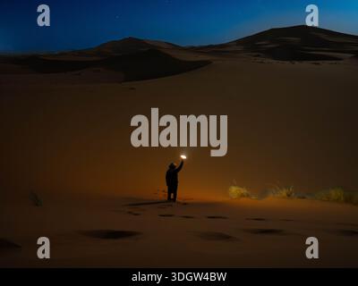 Solitary figure standing in complete darkness of the Moroccan Sahara, raising a flashlight that creates a small pool of light on the sand under a deep Stock Photo