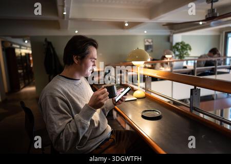 Relaxed thoughtful man drinking coffee and checking messages on smartphone in bookstore cafe space. Stock Photo