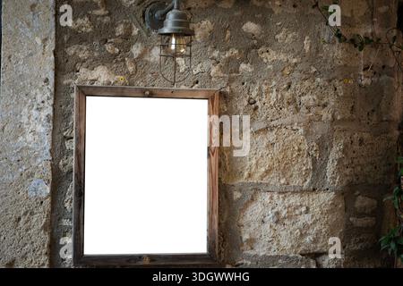 Blank wooden wooden frame with copy space hanging on a textured rustic stone wall, illuminated by a vintage wall lamp for a mockup display Stock Photo
