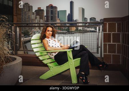 Woman Relaxing on Rooftop with Cityscape. A woman sits on a green chair on a rooftop overlooking a cityscape. She holds a book and appears relaxed in Stock Photo