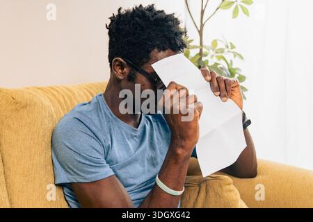 Young man sitting on a sofa at home covering his face with a bill, feeling stressed and overwhelmed by paperwork, expenses and financial responsibilit Stock Photo