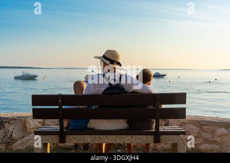 Back view of mother in straw hat with two sons sitting on bench looking at sea with boats at sunset. Concept of family vacation, Mediterranean travel, Stock Photo