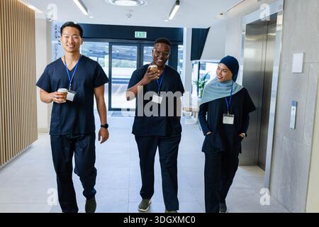 Diverse coworkers walking down hospital corridor wearing navy scrubs, carrying coffee cups, phone Stock Photo