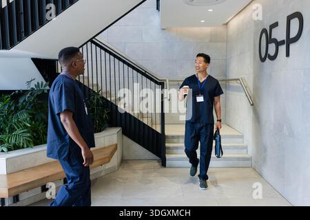 Diverse male coworkers in navy scrubs, blue IDs, chatting on stairs holding paper cup and bag Stock Photo