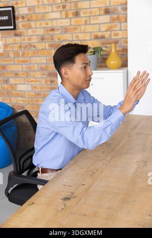 Asian man wearing blue shirt sitting at wooden table in workspace, gesturing with both hands Stock Photo