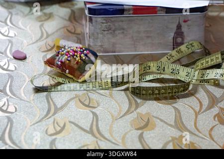 Colorful sewing workspace featuring a pincushion, measuring tape, glue, eraser, and British-themed tin box on patterned surface. Stock Photo
