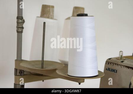 Johor, Malaysia, September 9, 2025: Three large white thread spools mounted on a stand attached to a JUKI sewing machine in a textile workspace. Stock Photo