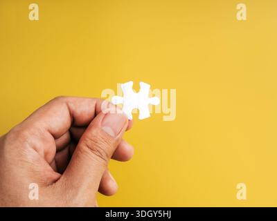 Hand holding a single white puzzle piece on yellow background symbolizes critical missing part, essential element, final step, problem solving, teamwo Stock Photo