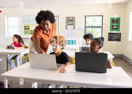 African teacher with school-age students leaning over desk assisting boy using laptop in classroom Stock Photo