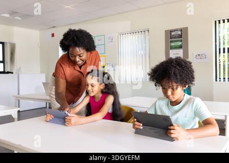 Diverse educator in rust-orange blouse guiding two youth using tablets at white table in classroom Stock Photo