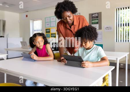 African American woman teacher leaning over table supervising two children using tablets in class Stock Photo