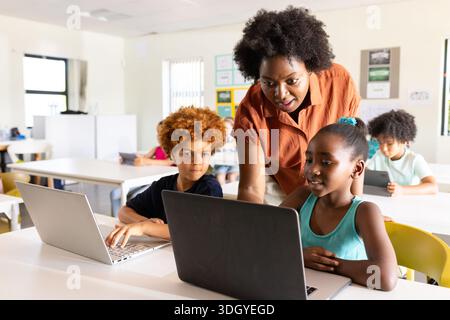 African American teacher wearing orange helping youth female in teal top using laptop in classroom Stock Photo