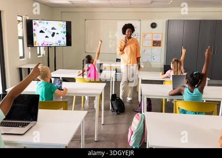 Woman teacher wearing orange shirt addressing students in classroom with laptops, whiteboard Stock Photo