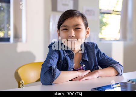 Female child sitting at bright classroom table wearing denim shirt, using tablet and notebook Stock Photo