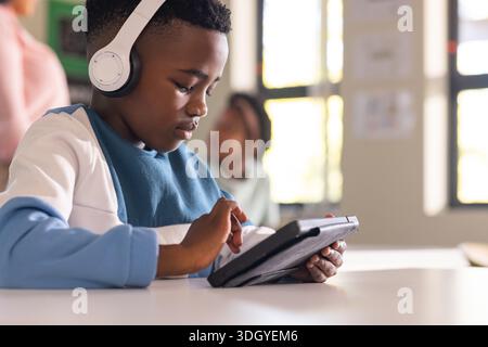 African American boy sitting in classroom at white table wearing white headphones and using tablet Stock Photo