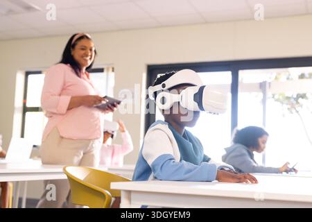 Teacher leading school pupils, boy sitting with white VR set, teacher holding tablet in classroom Stock Photo