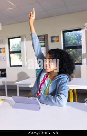 African American teen student sitting at white desk raising hand using purple tablet in classroom Stock Photo
