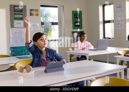 Diverse children in classroom sitting at desks raising hands while using tablet, laptop, headphones Stock Photo
