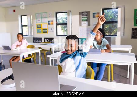 African American students sitting at classroom desks using laptop and tablets, boy raising hand Stock Photo