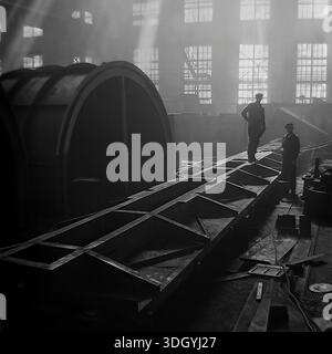 Slaviansk, Ukrainian SSR, Soviet Union, 1975. A dramatic black and white archival photograph taken inside the main assembly hall of the Slavtyazhmash heavy machinery plant. The image captures two workers standing near a massive steel chassis and a large cylindrical component, illuminated by shafts of sunlight streaming through the high factory windows. This atmospheric industrial scene illustrates the scale of heavy manufacturing and the daily labor of Soviet engineers building specialized railway wagons and metallurgical equipment in the peaceful Donbas region. Stock Photo