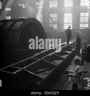 Slaviansk, Ukrainian SSR, Soviet Union, 1975. A dramatic black and white archival photograph taken inside the main assembly hall of the Slavtyazhmash heavy machinery plant. The image captures two workers standing near a massive steel chassis and a large cylindrical component, illuminated by shafts of sunlight streaming through the high factory windows. This atmospheric industrial scene illustrates the scale of heavy manufacturing and the daily labor of Soviet engineers building specialized railway wagons and metallurgical equipment in the peaceful Donbas region. Stock Photo