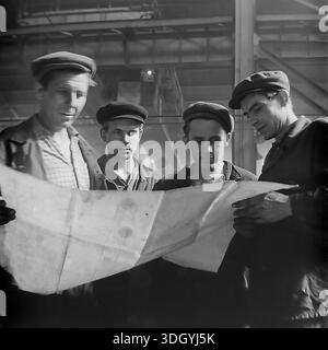 Slaviansk, Ukrainian SSR, Soviet Union, 1975. A compelling archival close-up photograph taken inside the Slavtyazhmash heavy machinery plant, featuring a team of four workers and engineers studying a large technical blueprint. Dressed in traditional flat caps and work jackets, the men are deeply focused on the engineering drawing, holding it open together. This detailed group portrait highlights the importance of intellectual labor, collaboration, and precise planning in the manufacturing industry of the peaceful Donbas region. Stock Photo