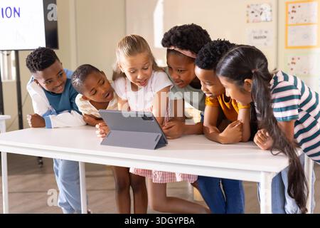 Diverse group of six school-age children gathering around tablet in gray folio case at class table Stock Photo