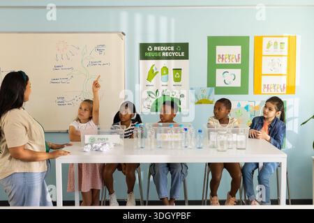 Diverse teacher with schoolchildren pointing to whiteboard in classroom with plastic bottles Stock Photo