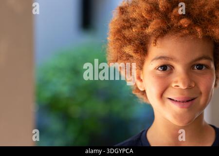 School-age African American boy smiling on porch in dark crew-neck shirt near green foliage Stock Photo