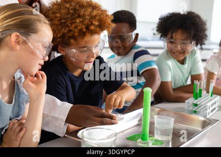 Diverse goggled students lab-coated woman teaching at lab watching green foam rising from cylinder Stock Photo