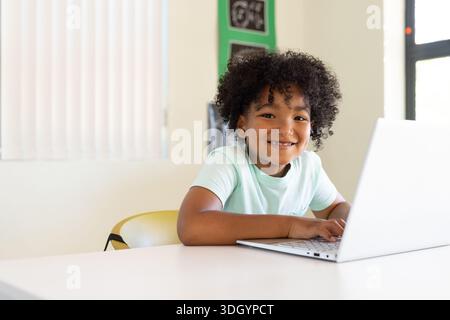 African American schoolboy sitting at white table in classroom typing on silver laptop in mint tee Stock Photo