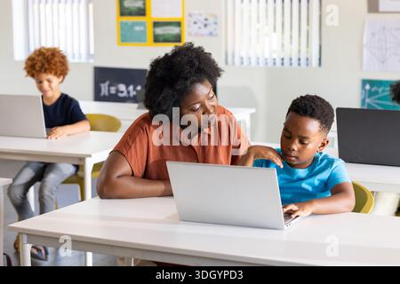 Female teacher in rust blouse leaning toward youth student guiding on silver laptop in class Stock Photo