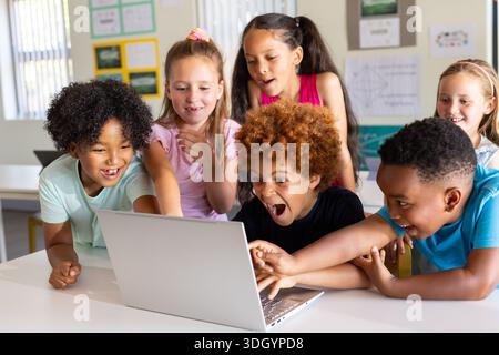 Diverse children leaning in around silver laptop on white table in classroom, pointing and reaching Stock Photo
