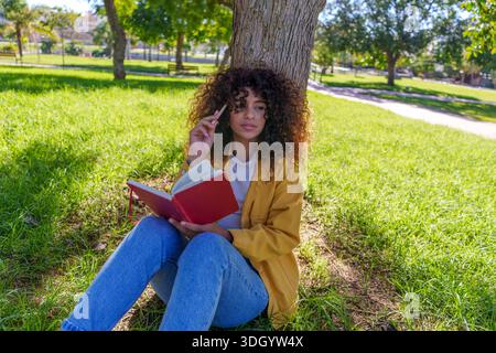 Woman with curly hair sitting on grass leaning on a tree trunk, holding a red notebook and pencil, thinking and creating Stock Photo