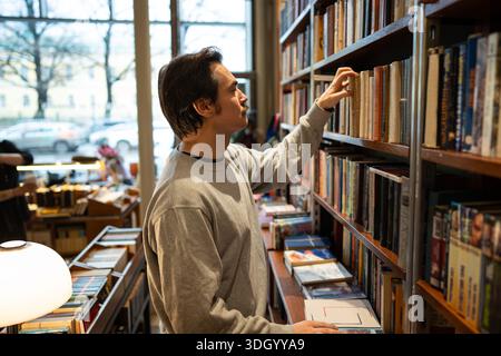Curious focused man booklover picking hardcover book from bookshelf in modern bookstore library. Stock Photo