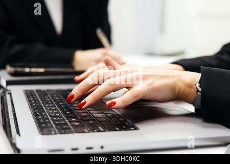 Businesswoman with red nails is typing on a laptop at a desk, blurred colleague in the background holding a pen during a collaborative work environmen Stock Photo
