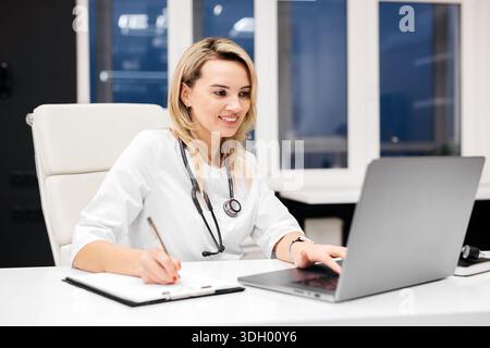 Female doctor is engaged with a laptop at a sleek office desk, taking notes on a clipboard with large windows allowing natural light in Stock Photo