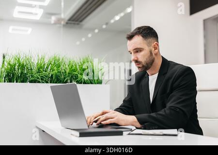 Businessman in a black suit is focused on his laptop at a modern office desk, with green plants providing a fresh touch in the background Stock Photo