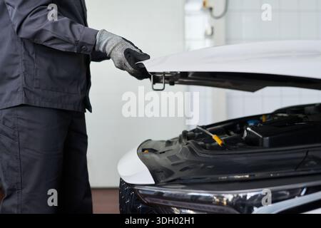 Anonymous man wearing work gloves opening car hood in garage, preparing for vehicle inspection or maintenance, only upper body and hands visible, focus on action Stock Photo