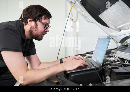 Caucasian young adult man working on car diagnostics using laptop computer under open hood in auto repair shop, focusing on analyzing engine performance and troubleshooting issues Stock Photo