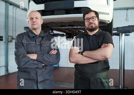 Portrait of two middle aged Caucasian men standing with arms crossed in auto repair shop, looking confidently at camera, car lifted on hydraulic platform in background Stock Photo