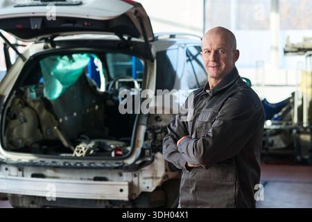 Portrait of middle aged Caucasian man standing with arms crossed in front of disassembled car in auto repair shop, looking at camera with confident expression Stock Photo