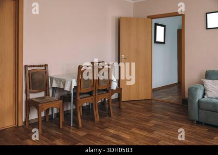 A cozy hotel room with wooden flooring. A small dining table with four chairs is set against a light pink wall. A door leads to another room, and a so Stock Photo