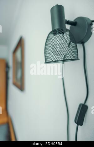 A modern wall lamp with a mesh shade is mounted on a white wall. The lamp features a black cord and a switch, adding a stylish touch to the hotel room Stock Photo