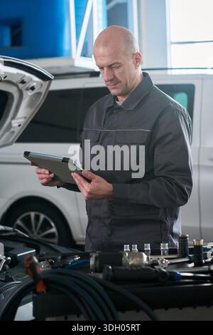 Middle aged Caucasian man standing in auto repair shop using digital tablet while inspecting car engine, focusing on diagnostics and maintenance tasks in professional environment Stock Photo