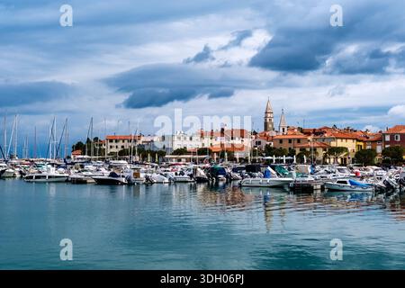 Small boats moored in a harbor with a town and the bell towers of two churches in the distance under a cloudy sky. Stock Photo