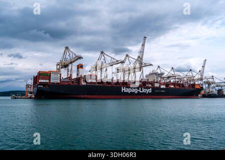 Large Hapag-Lloyd container ship docked at a commercial port terminal with several loading cranes under a cloudy sky. Stock Photo