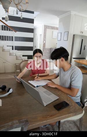 Asian students studying together at open-plan kitchen table using laptop tablet notebook headphones Stock Photo